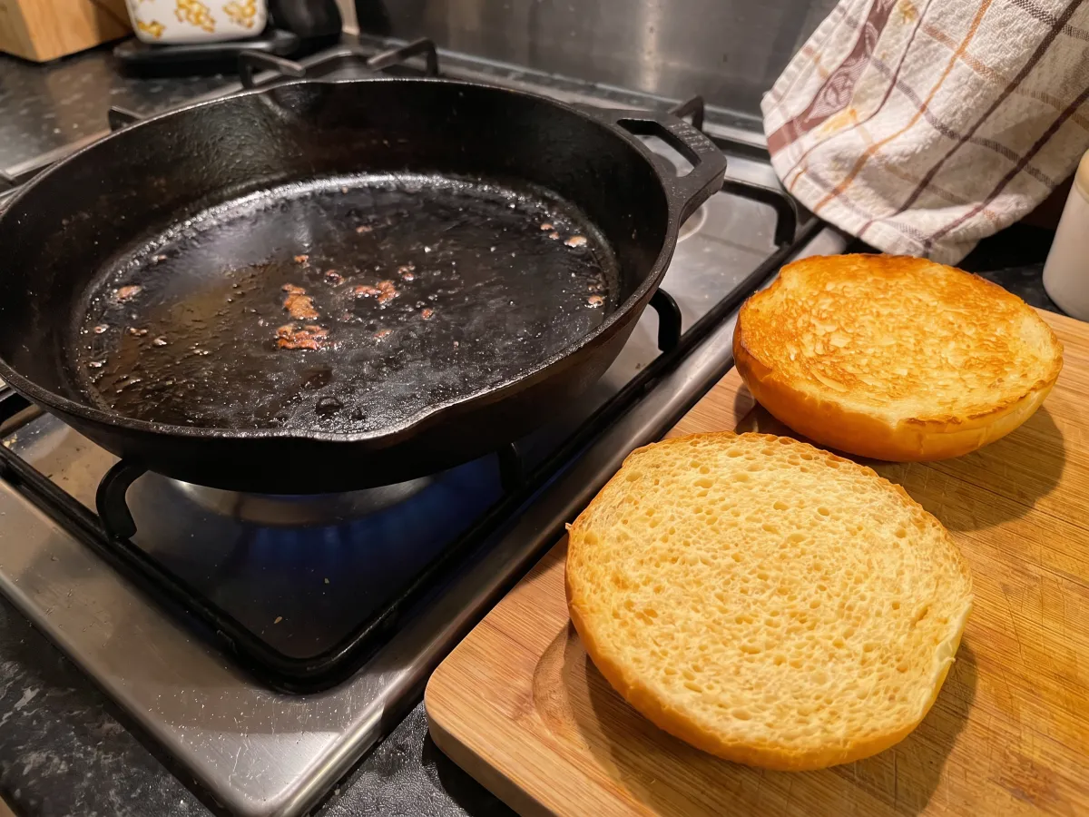 Pão brioche aberto junto à frigideira da carne: uma metade sem tostar e a outra tostada