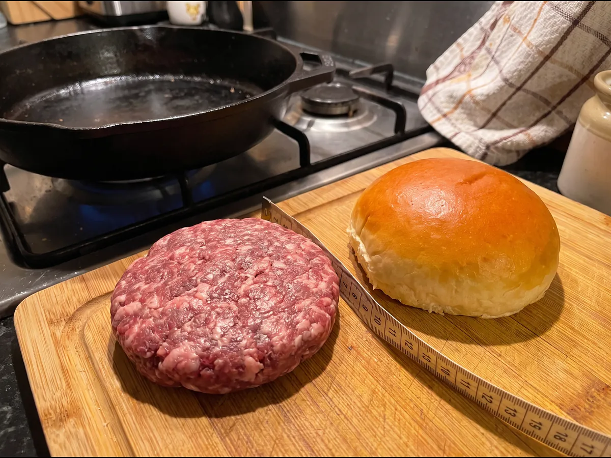 Um pão de hambúrguer brioche ao lado de um hambúrguer cru um pouco mais largo, com uma fita métrica ao lado