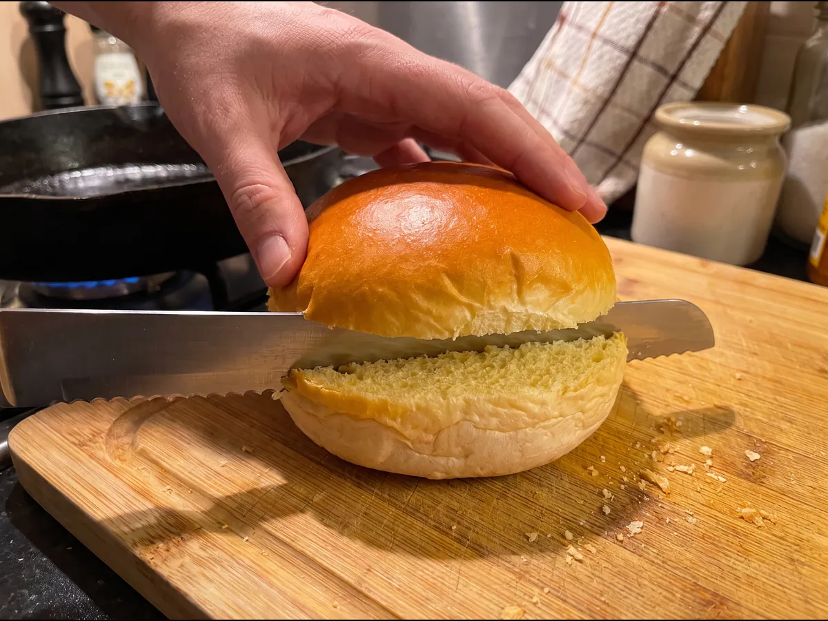 Um pão brioche a ser aberto na horizontal com uma faca de serrilha numa tábua